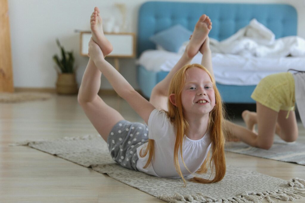 Full body of cheerful girl in casual clothing with reddish hair looking away with smile while stretching legs on carpet in bright cozy living room on blurred background